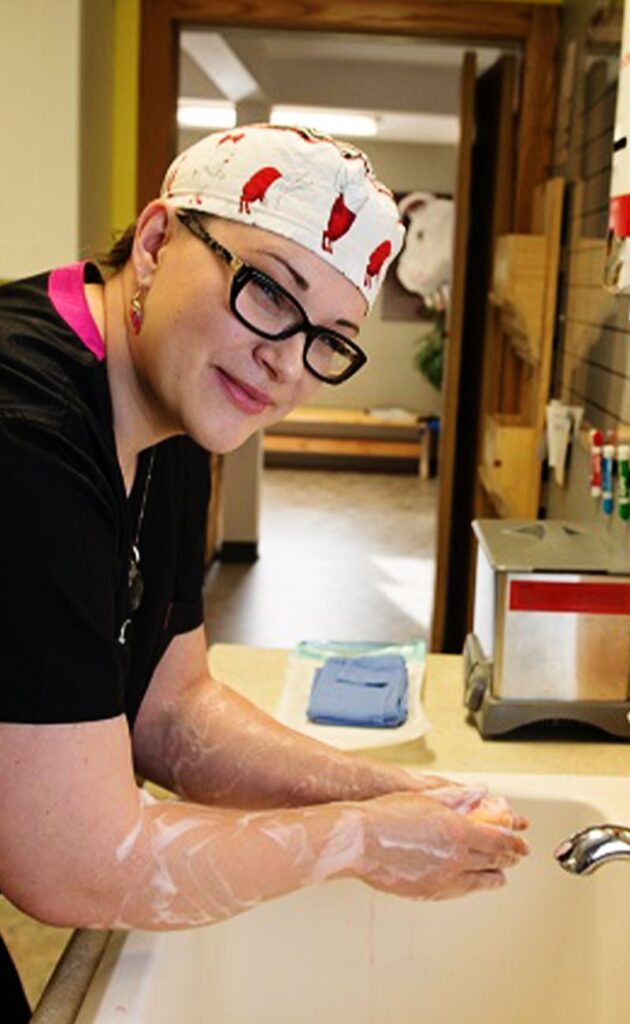Woman in glasses washing her arm at a sink, wearing a bird patterned cap.